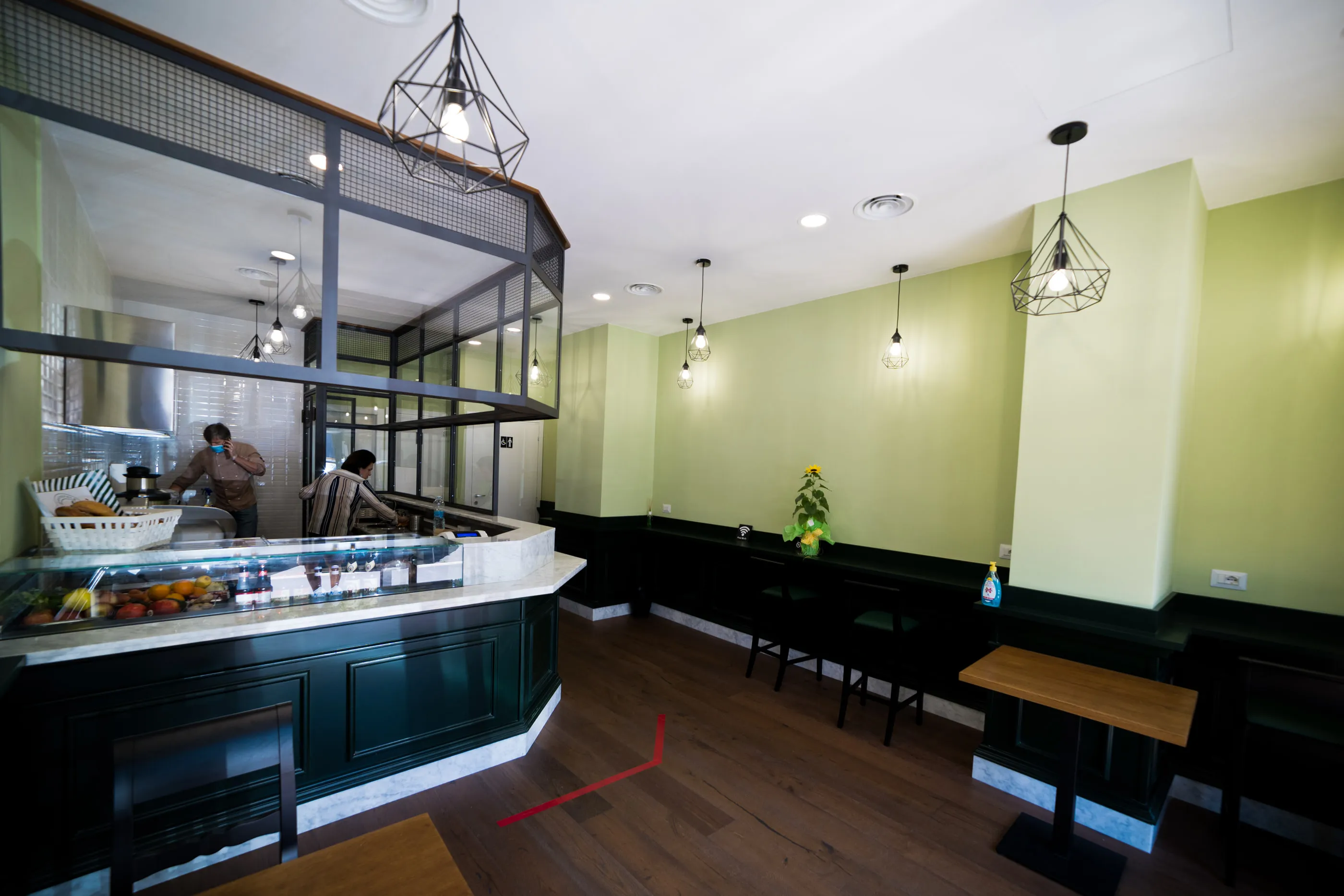 Sala di una caffetteria con pareti verde salvia, banco in legno laccato scuro e lampade a sospensione in metallo nero