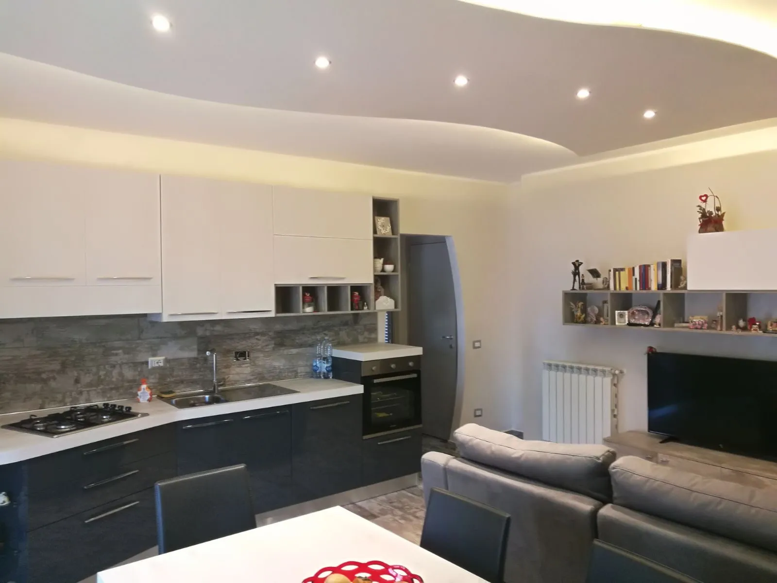 Soggiorno con cucina a vista, ante blu e bianche e controsoffitto curvo con faretti incassati